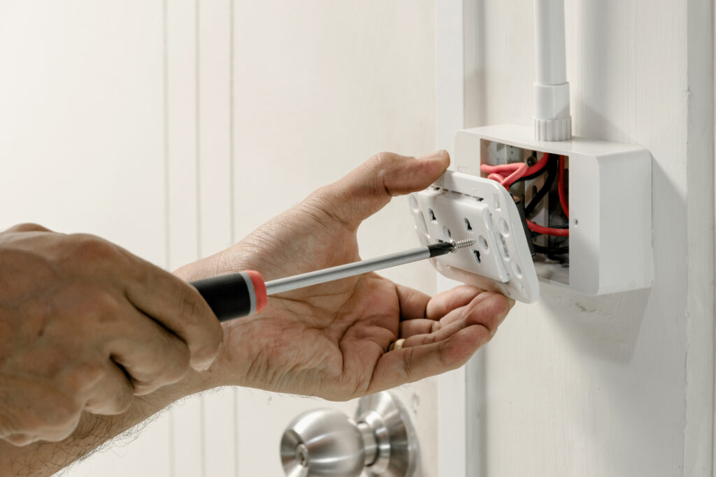 An electrician is using a screwdriver to attach a power cord to a wall outlet.